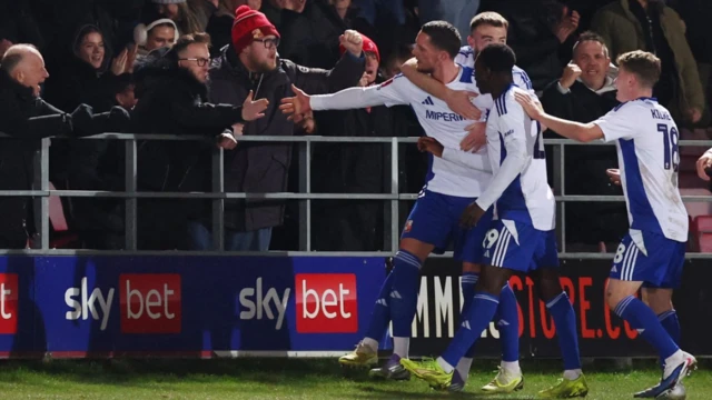 Cuatro jugadores del Swindon Town vestidos de azul y blanco se disponen a estrechar la mano de los aficionados que se encuentran junto a las barandillas del campo.