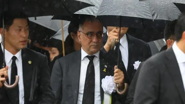 黒いスーツと黒いネクタイを締め、胸に白い胸花をつけ、黒い傘を持ったイスラエル大使