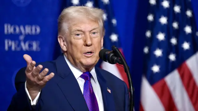 ترامب يتحدث خلال اجتماع مجلس السلام في واشنطن العاصمة في 19 فبراير/شباط 2026. وهو يرفع يده اليمنى أثناء حديثه عبر الميكروفون، والعلم الأمريكي خلفه.