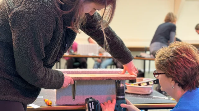 Dos mujeres instalan un estante de madera artificial sobre un bloque con un taladro. Una de ellas sostiene el estante sobre soportes mientras la otra usa el taladro para instalarlo. Se ve a Sarah Bolger al fondo hablando con otra persona.