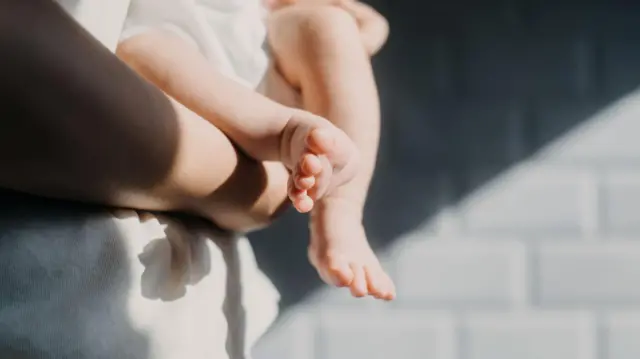 Tampilan jarak dekat dari kaki bayi. Bayi itu digendong oleh orang dewasa yang mengenakan kemeja putih, berdiri di depan dinding bata abu-abu
