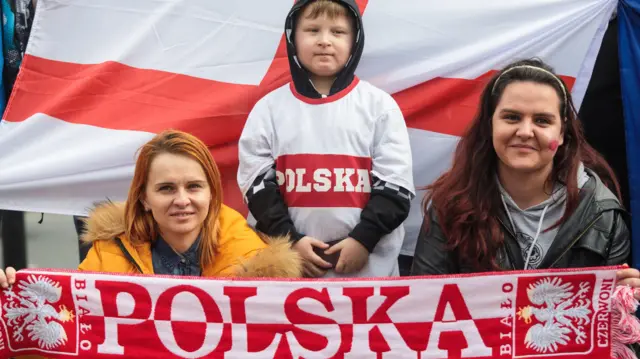 Dwie kobiety i chłopiec stoją przed angielską flagą, trzymając szalik z napisem Polska. Chłopiec ma na sobie koszulkę z polską flagą.