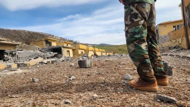 Зруйновані будівлі з жовтими стінами, які частково обвалилися. На передньому плані стоїть людина у камуфляжному одязі та черевиках, дивлячись у бік руїн. Довкола – уламки бетону та сміття, на горизонті – пагорби під ясним небом.