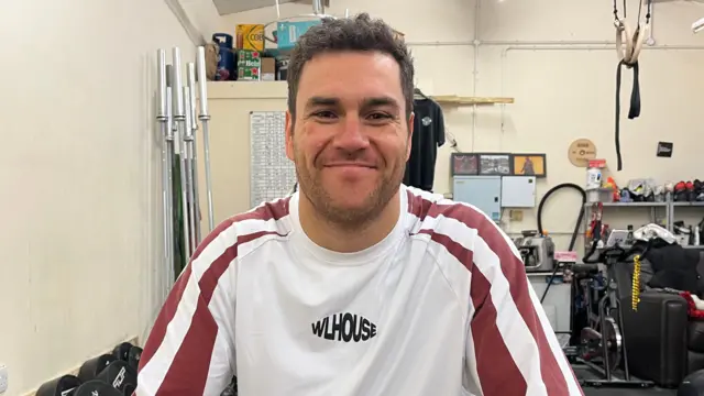 El Sr. Heenan aparece en la foto dentro del gimnasio, mirando a la cámara y sonriendo. Lleva una camiseta de rayas blancas y rojas. El gimnasio tiene paredes blancas y una hilera de pesas apiladas en una esquina, con pequeñas mancuernas negras apiladas junto a ellas. Sobre su hombro hay una estantería metálica con varios aparatos de gimnasio.