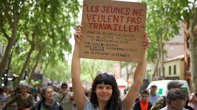 O tânără ține în mână o pancartă pe care scrie: „Tinerii nu vor să muncească pe un salariu de subzistență, pentru un sistem capitalist și neocolonial, fără drepturi la șomaj și pensie, în timp ce planeta este distrusă.”