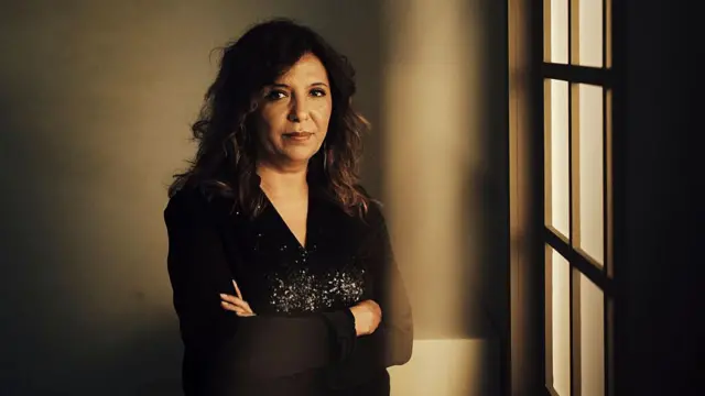 Ben Hania siyah bir kıyafetle bir pencerenin önünde ayakta duruyor. Ellerini önünde birleştirmiş. 