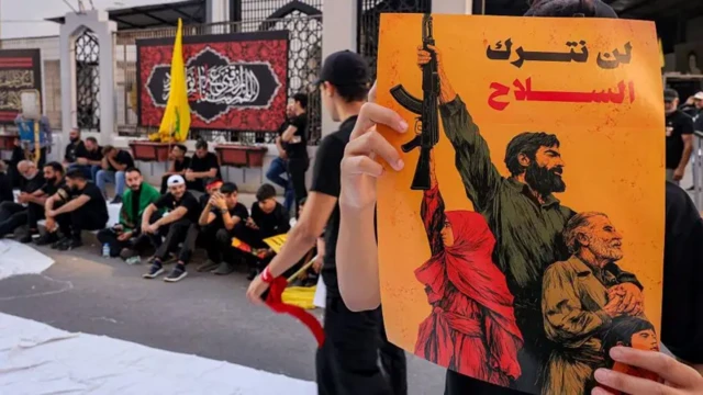 تصویری از نقاشی یک جوان مسلح که دختری جوان، مردی سالخورده و کودکی او را احاطه کردهاند. دو دست این نقاشی را نگه داشتهاند و روی آن نوشته شده است: «سلاح را کنار نخواهیم گذاشت.» حامیان حزبالله با لباس سیاه بر لبه خیابان نشستهاند