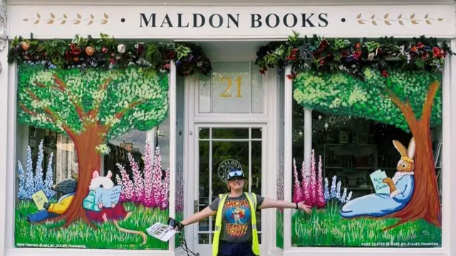 Una tienda tiene un letrero blanco en la parte superior que dice "Libros Maldon" en letra negra. Los escaparates están llenos de cuadros de árboles, flores y criaturas del bosque leyendo libros. Una mujer con un top reflectante, camiseta, gorra y gafas de sol se encuentra de pie con los brazos en alto fuera de la tienda.