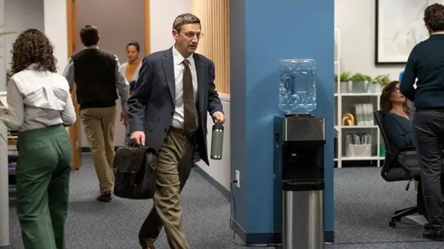 Кадр з серіала "Між крісел". На фото - офіс з багатьма людьми. В центрі - чоловік, який проходить повз кулер