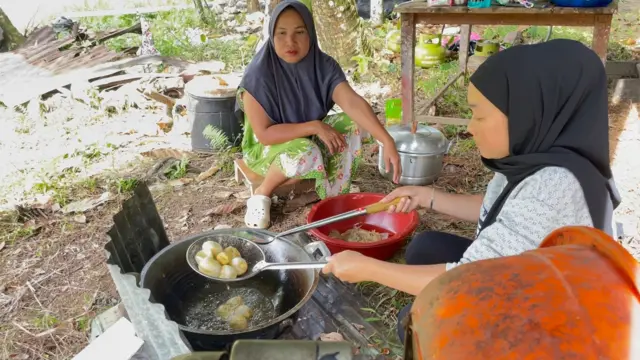 Seorang perempuan berkerudung hita, sedang mengangkat telur bulat yang baru selesai digoreng, sementara satu perempuan lagi berkerudung abu-abu dan memakai daster hijau, memperhatikan di sebelahnya. Mereka sedang berada di dapur umum di lokasi huntara.