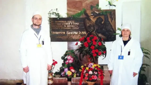 Чоловік і жінка у білих халатах та шапочках стоять по обидва боки меморіальної плити, уквітчаної великими букетами червоних, рожевих і жовтих квітів. На плиті – рельєфна скульптурна композиція та напис українською мовою: "Не залишили пости, мужньо стояли у герці, памʼятник їм вознести треба у кожному серці. Ходимчук Валерій Ілліч"