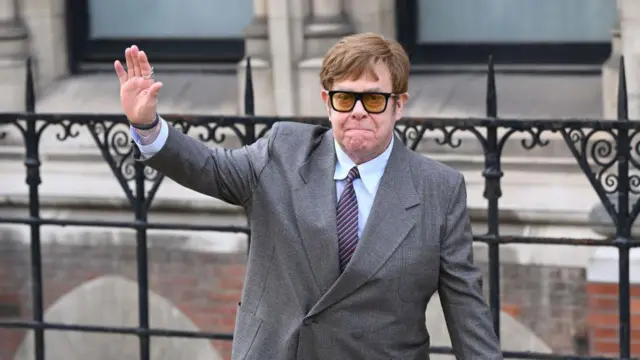 Sir Elton John in a suit, tie and sunglasses, waving outside the Royal Courts of Justice in 2023