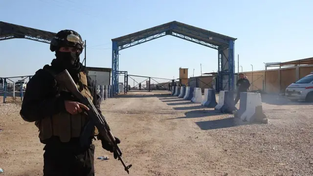 Açık hava cezaevinin girişinde bir Suriyeli silahlı kişi kameraya bakıyor. Toprak alanda arkada giriş kapısı var.