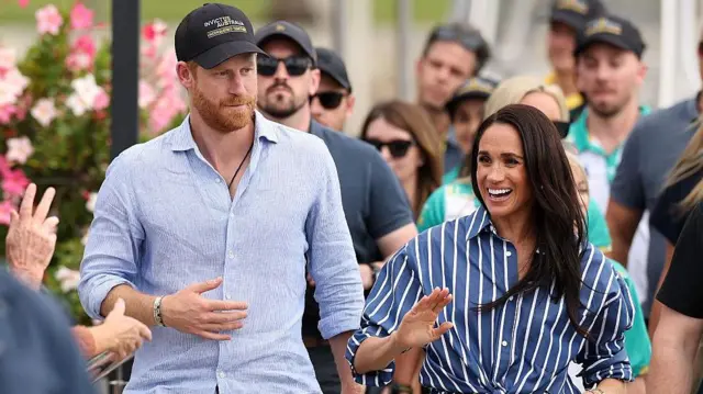 Ducele și Ducesa de Sussex merg pe jos. Harry poartă o cămașă albastră și o șapcă de baseball bleumarin, iar Meghan poartă o cămașă cu dungi albastre și albe. Ea are părul închis la culoare, purtat desfăcut.