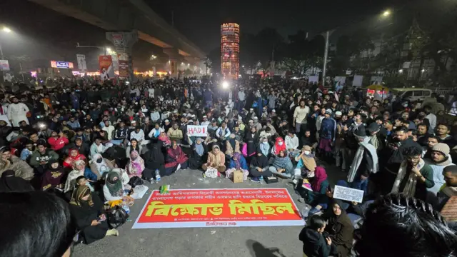 শুক্রবার রাতে শাহবাগে ইনকিলাব মঞ্চের অবস্থান কর্মসূচিতে অংশ নিয়ে অনেকে বসে ও দাঁড়িয়ে আছেন। বিক্ষোভ মিছিল লেখা একটি ব্যানার মাঝখানে বিছানো আছে