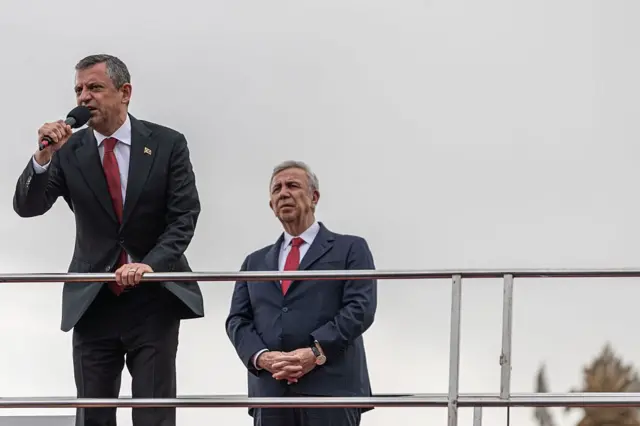 Bir otobüsün üzerinde takım elbiseli iki adam. Biri önde mikrofonda konuşuyor. Diğeri arkada ellerini kovuşturmuş.