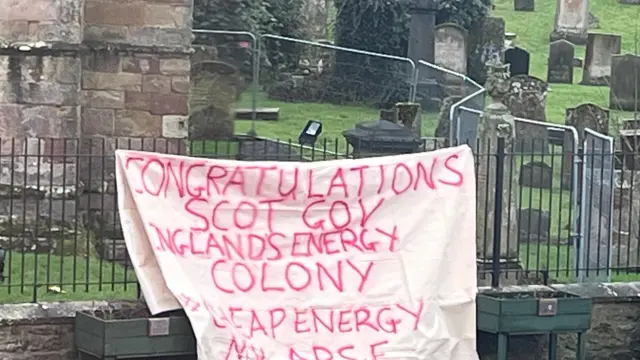 Una pancarta blanca con letras rojas que dice "Felicitaciones al gobernador escocés, Colonia Energética de Inglaterra" cuelga de una barandilla frente a un cementerio.