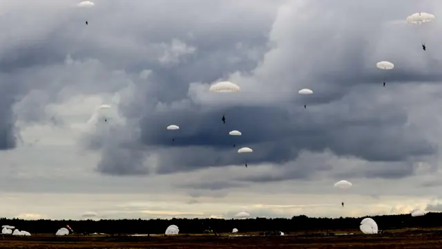 Około tuzina żołnierzy ze spadochronami na tle ciemnego, zachmurzonego nieba. Kilku żołnierzy już wylądowało.