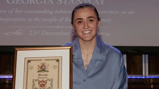 Georgia Stanway, de cabello castaño, con una blusa azul y un collar, sonríe y sostiene un certificado que le otorga el premio "Libertad del Pueblo".
