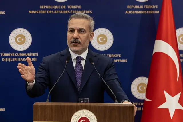 Dışişleri Bakanı Hakan Fidan tek eli havada kürsünün arkasından açıklama yapıyor. Fotoğrafın sağ tarafında Türk bayrağı var. 