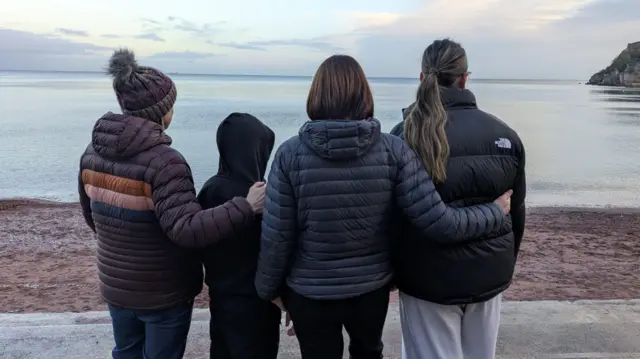 Laura, James, Beth i Kate stoją obok siebie tyłem do kamery, patrząc na spokojne morze z linii brzegowej.