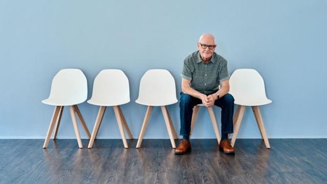 Homem de cabelo branco sentado aguardando ao lado de cadeiras vazias