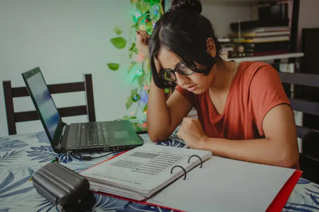 Mujer con gafas en una mesa leyendo unos apuntes de un archivador, con una computadora al lado.