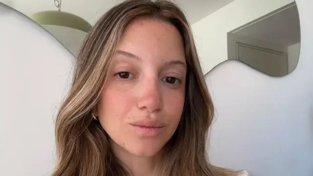 في هذه الصورة الذاتية، تنظر مورا إلى الكاميرا بابتسامة. ينسدل شعرها، الممزوج بخصلات شقراء، على كتفيها. ترتدي أقراطاً ذهبية صغيرة.