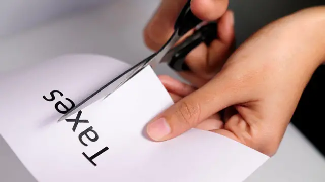 Sebuah gambar kreatif memperlihatkan tangan yang sedang memotong tulisan "pajak" pada selembar kertas menggunakan gunting.