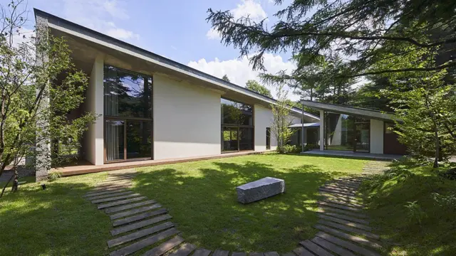 Casa moderna de una planta rodeada de árboles, con grandes ventanales, jardín de césped y senderos de madera curvos.