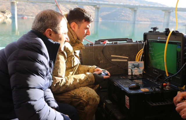 Bir asker ve sivil yetkili bir ekrana bakarak deniz altındaki yapılan aramaya ilişkin görüntüleri izliyor. Arkada bir köprü var.