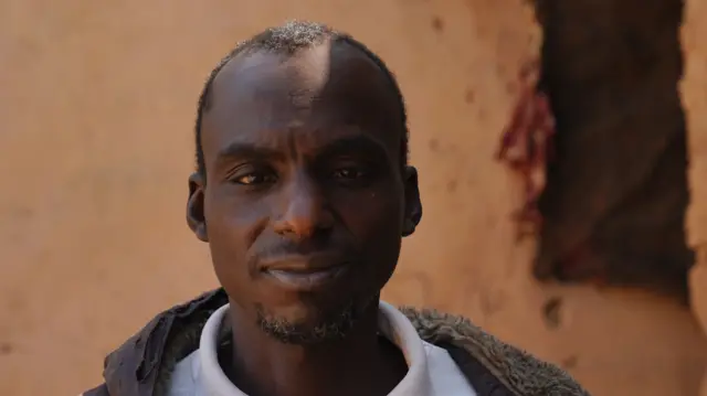 Idris Alhassan, vêtu d'un tee-shirt blanc, regarde la scène en plissant les sourcils et en se tapotant l'épaule avec sa veste grise.