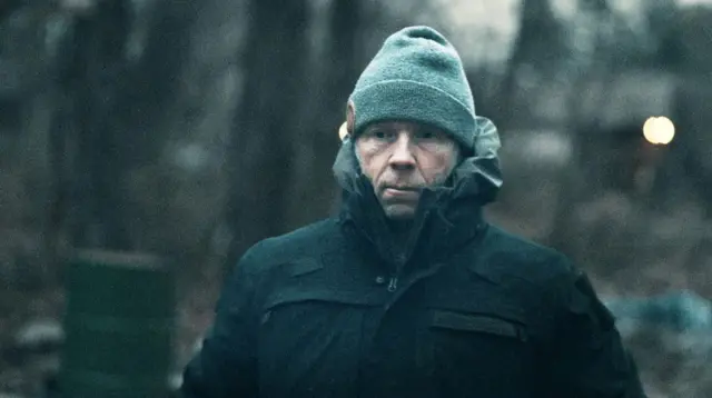Грег Сквайр — мужчина в шапке и тёмно-зелёной куртке с капюшоном стоит на улице в пасмурную погоду. Его лицо серьёзное, взгляд направлен в камеру, на заднем плане видны деревья без листвы