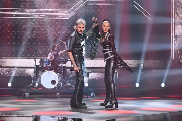 мушкарац и жена обучени у кожне панталоне и кошуље држе се за руке и гледају у камеру. иза њих је бубњар за бубњевима, држи палице у вис