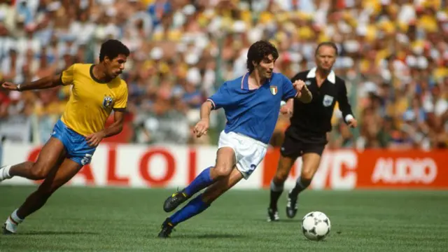 A imagem mostra um jogador da seleção italiana conduzindo a bola em velocidade durante uma partida contra o Brasil, enquanto é perseguido por um defensor brasileiro. Ao fundo, o árbitro acompanha o lance e as arquibancadas lotadas reforçam o clima de uma decisão em um grande estádio.