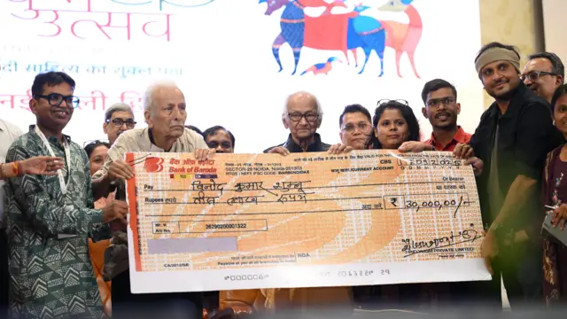  विनोद कुमार शुक्ल और अन्य एक चेक का नमूना पकड़े हुए 