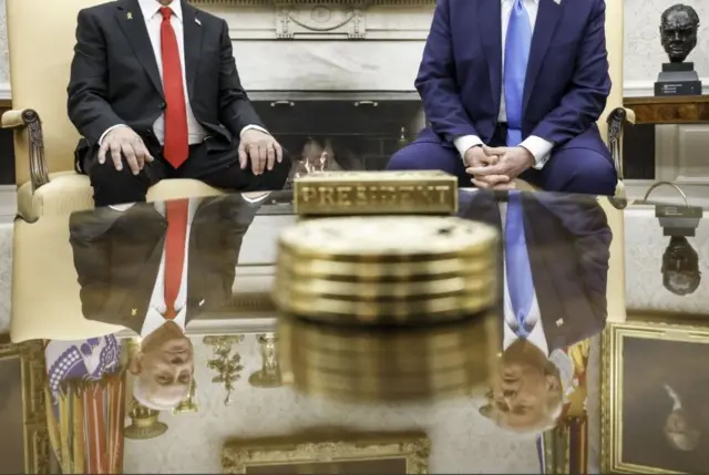 رئيس الوزراء الإسرائيلي بنيامين نتنياهو (يسار) بلباس رسمي أسود، والرئيس الأمريكي دونالد ترامب (يمين) بلباس رسمي كحلي، يتحدثان مع الصحفيين في المكتب البيضاوي بالبيت الأبيض. الصورة أخذت بطريقة مختلفة بحيث يظهر وجه ترامب ونتنياهو على انعكاس الطاولة أمامهما. 