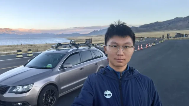 یوکون تائو در کنار جاده با خودرویش که خراب شده در پسزمینه دریاچه‌ای دیده می‌شود