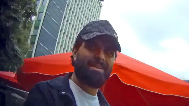 Un homme barbu aux cheveux noirs portant une casquette de baseball Gucci, un t-shirt blanc et une chemise bleu marine, regarde le journaliste sous couverture. En arrière-plan, on aperçoit un parasol de café orange et un gratte-ciel.