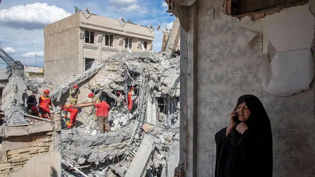 27 Mart 2026'nın erken saatlerinde Tahran'da hava saldırısında hasar gören bir konut binasının enkazında acil durum ekipleri arama yaparken bir kadın cep telefonuyla konuşuyor.