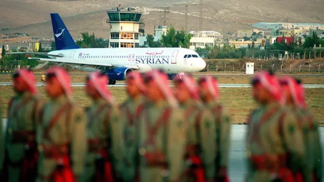 طائرة الرئيس السوري بشار الأسد تصل إلى مطار عمان، كما تظهر من خلف حرس الشرف الأردني في 18 أكتوبر 2000 في عمان، الأردن. وصل بشار الأسد إلى الأردن في أول زيارة عمل له إلى مملكة الملك عبد الله الثاني منذ انتخابه في يوليو لمناقشة العنف في الشرق الأوسط. 