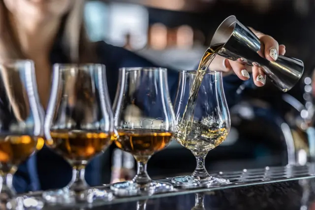 Jovem barman profissionalmente serve rum ou conhaque de um medidor de aço em um copo no bar.
