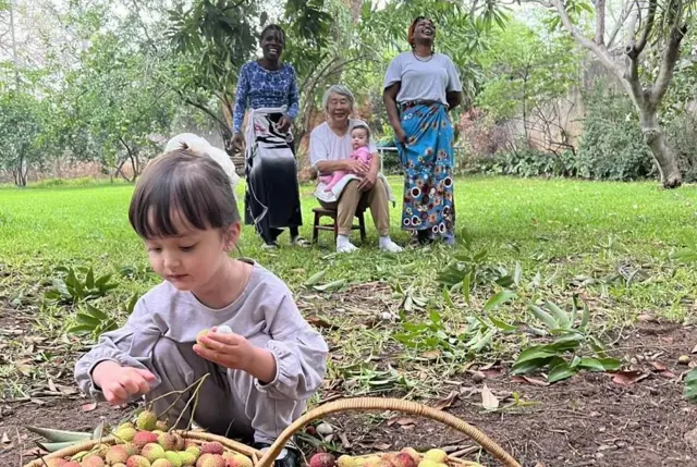 Дівчинка сидить перед кошиком з фруктами, на задньому фоні - три жінки, одна азійського походження, інші дві африканки, в азійської жінки на руках маленька дитина 