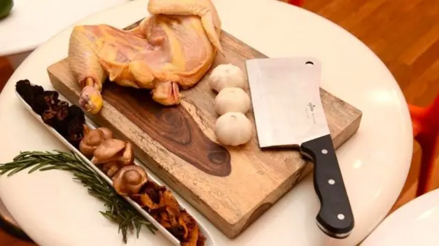 bir kesme tahtasının üzerinde çiğ tavuk budu, üç baş sarımsak ve büyük bir bıçak 