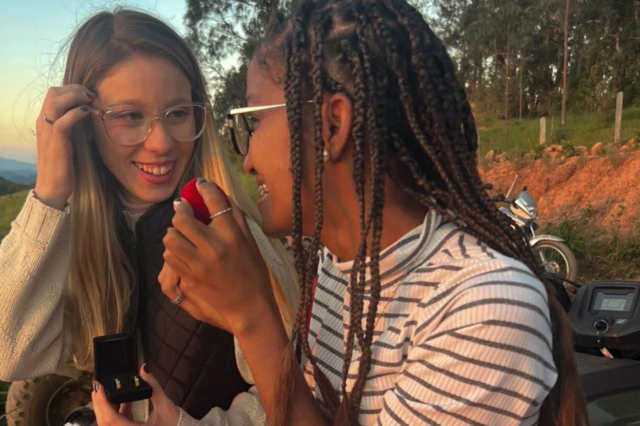 Fotografia colorida mostra duas mulheres sorrindo sentadas ao livre. Elas seguram caixinhas de anel de noivado e olham uma para outra