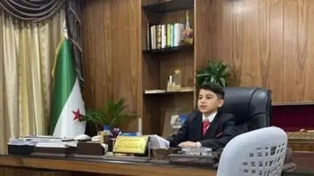 پسری با کت و شلوار و کراوات پشت میزی نشسته است