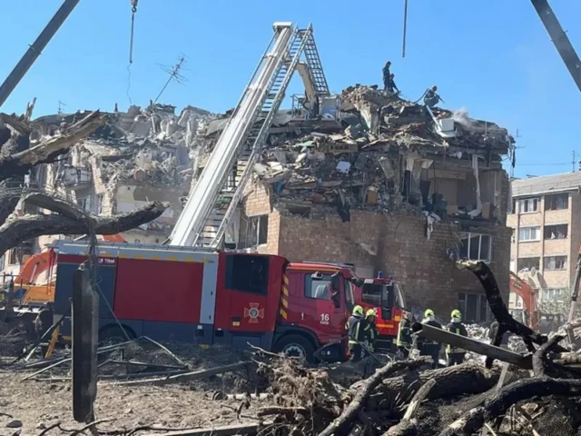 У развалин дома стоит пожарная машина и пожарные, лестница пожарной машины лежит на том, что осталось от крыши и верхних этажей, и там поверху тоже ходят люди, вероятно, спасатели.