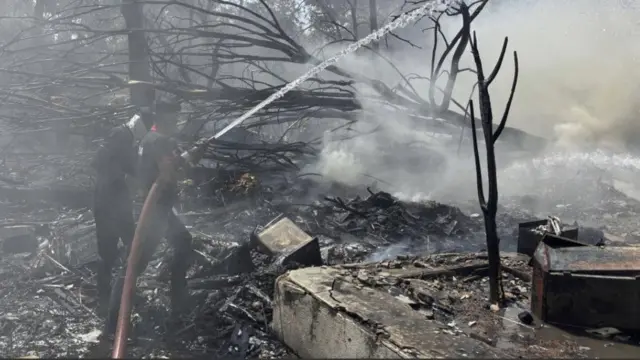 黒く焼け焦げて倒れた街路樹とがれきの中で、消防隊員がホースから放水している