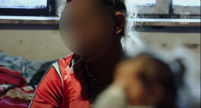 Kobieta i dziecko siedzą na posłaniu, pod oknem. Ich twarze zostały rozmyte