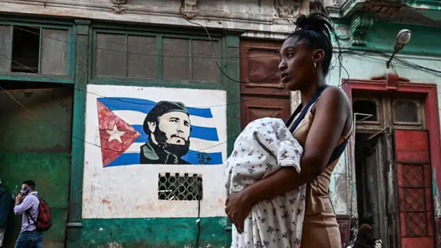 Arkada küba bayrağı üzerinde Fidel Kastro resmi çizili olan bir duvar var. Önde bir kadın elinde bir bebekle yürüyor. 
